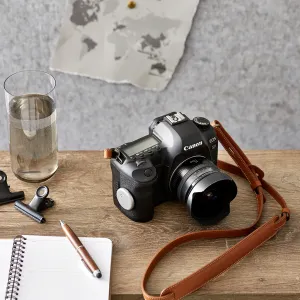 Cámara DSLR Canon EOS 5D con objetivo y correa marrón sobre una mesa de madera, junto a un cuaderno y un vaso de agua