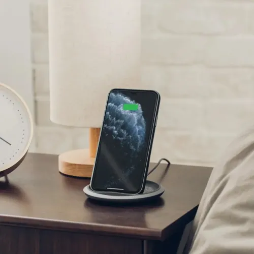 Moshi Lounge Q belaidis įkroviklis, laikantis išmanųjį telefoną vertikaliai ant naktinio staliuko, su įkrovimo indikatoriumi.